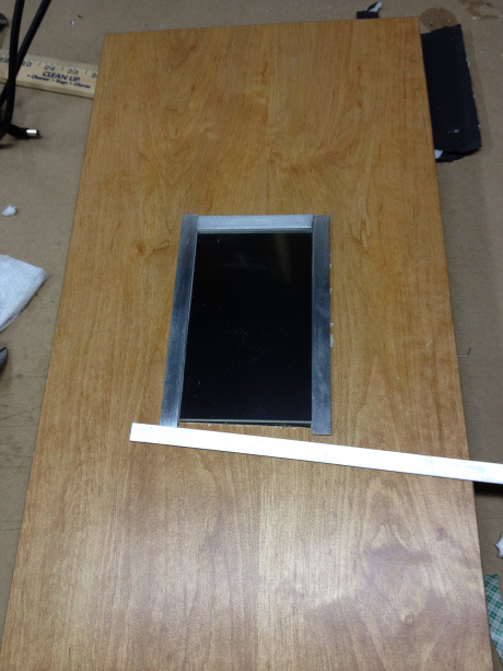 A rectangular cutout in a plywood panel with the screen dropped into it
