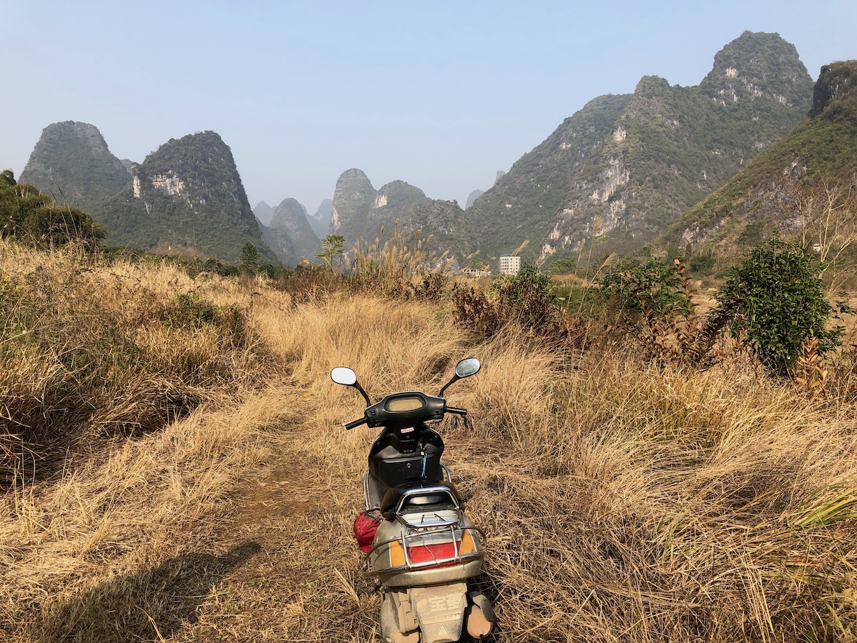 A scooter parked on a grassy trail leading toward karst towers outside Yangshuo