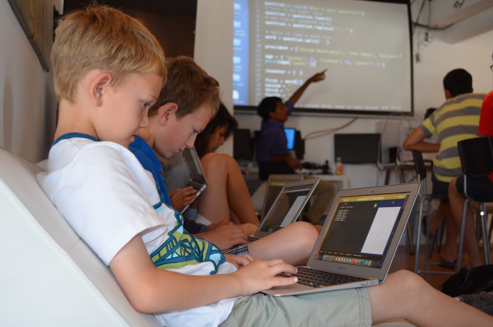 Students coding on laptops in a classroom while an instructor points at code projected on a screen