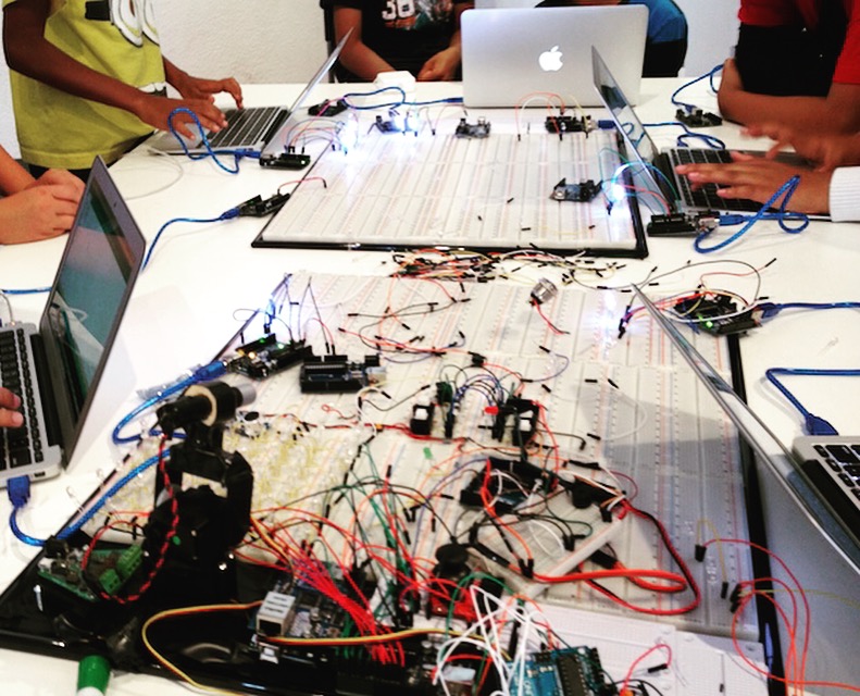 Students working on laptops around a table covered in breadboards and wiring